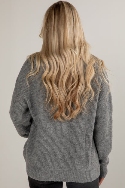 Person wearing a gray sweater with long blonde hair, standing against a plain background