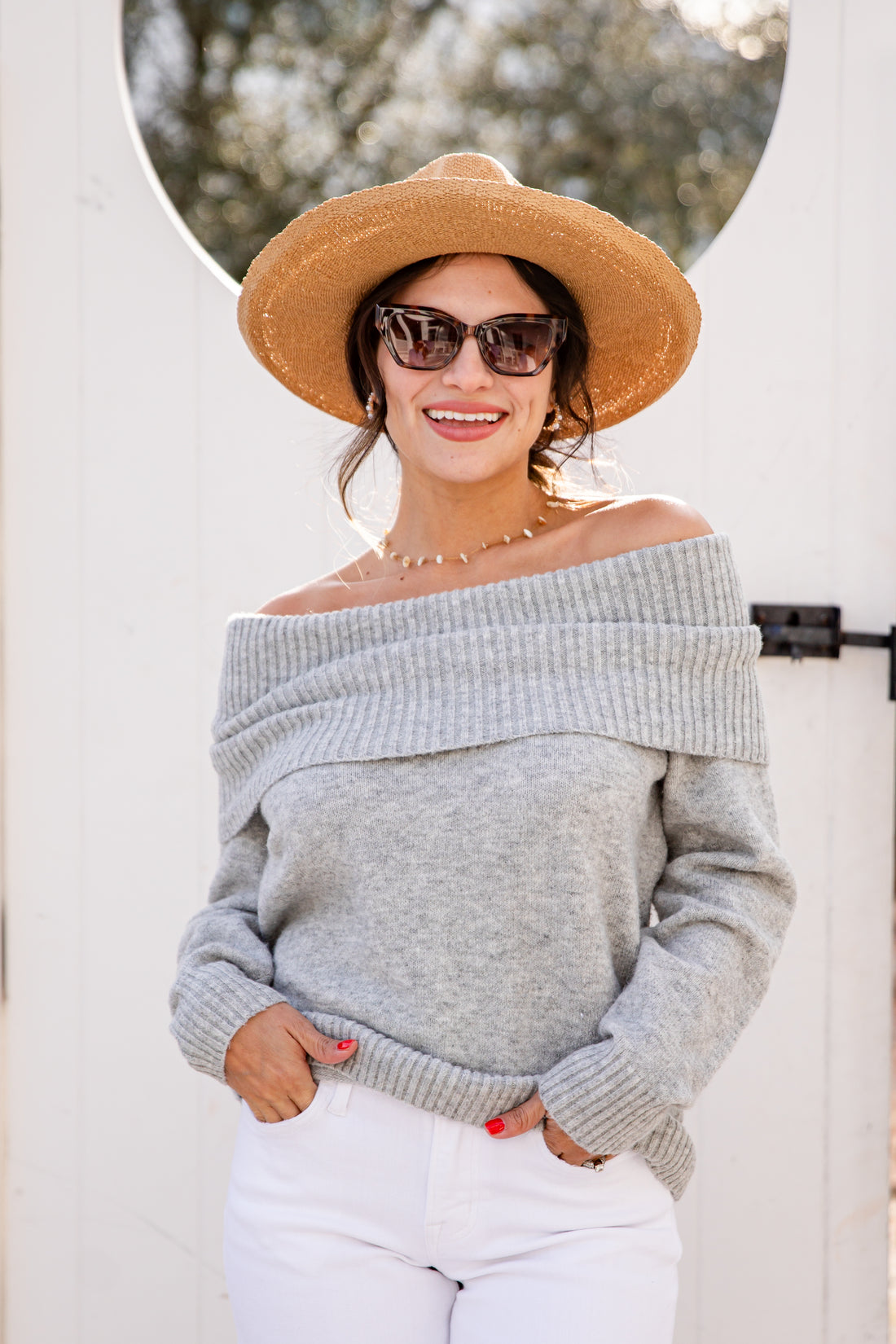 Woman wearing a gray off-shoulder sweater, white pants, and a straw hat outdoors.