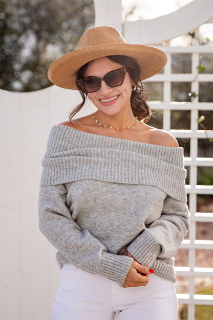 Woman wearing a gray off-shoulder sweater, white pants, and a brown hat with sunglasses.