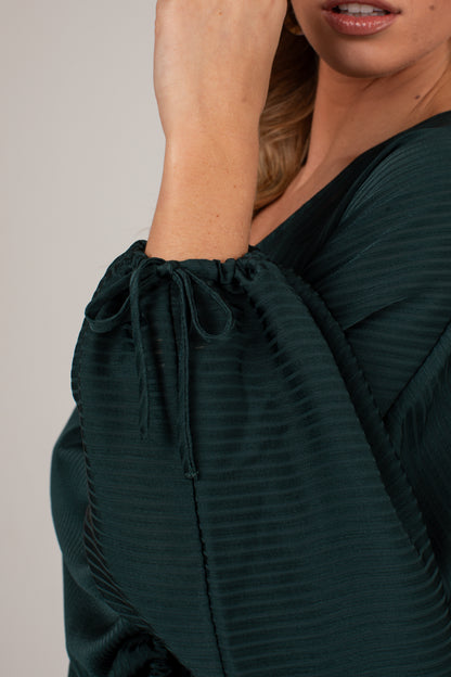 Close-up of a woman wearing a dark forest green pleated top with a drawstring puff sleeve, against a neutral light gray background.