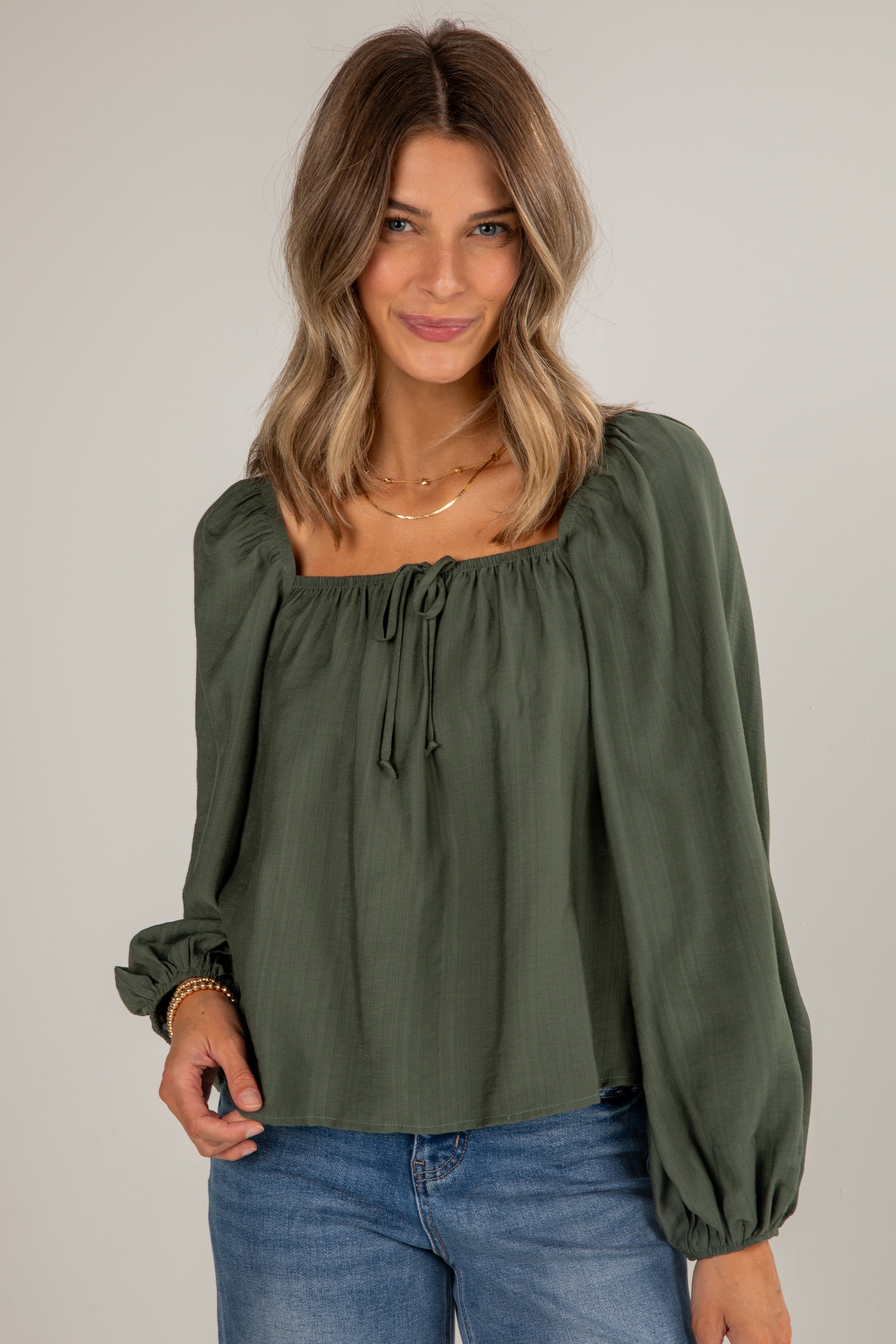 Woman wearing an olive green blouse with long sleeves and blue jeans on a plain background