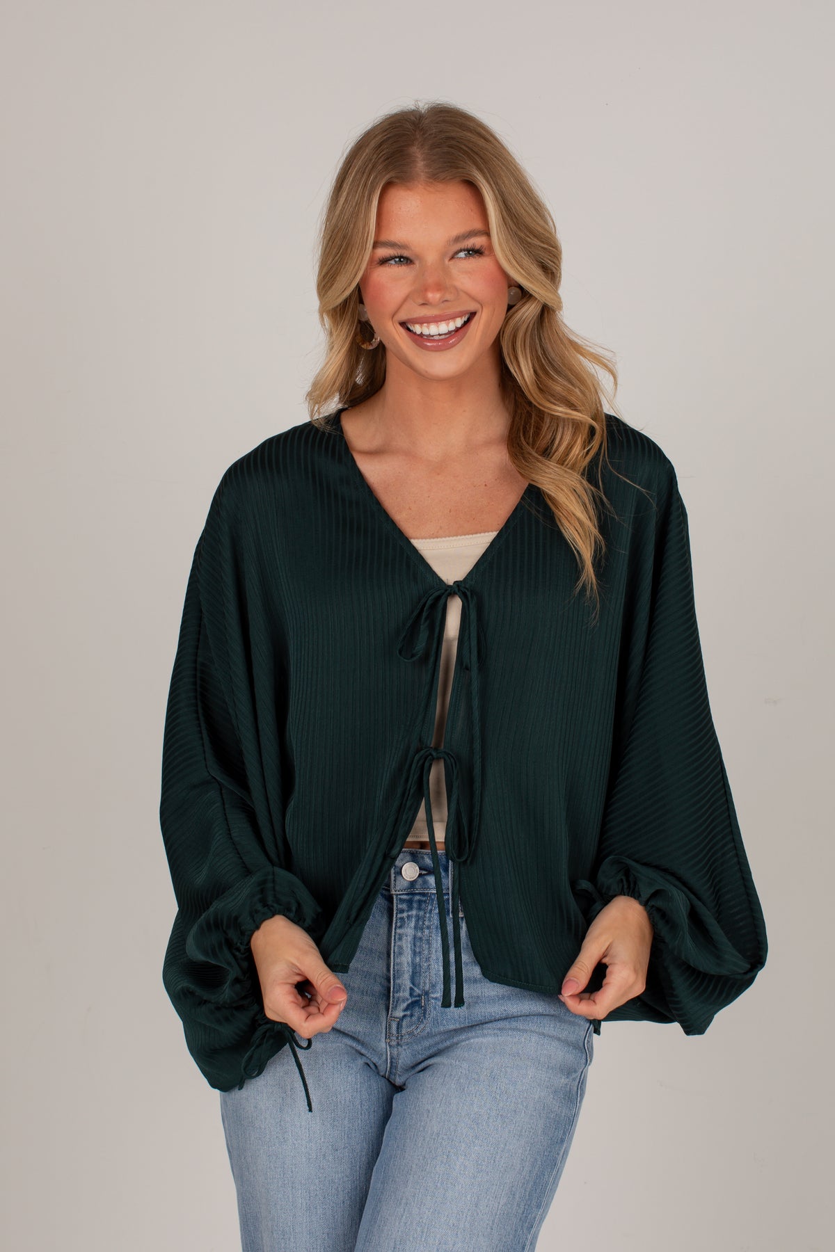 A smiling blonde woman wearing a dark forest green textured long-sleeve wrap top with a tie-front detail, styled with light-wash blue jeans against a plain light gray studio background.
