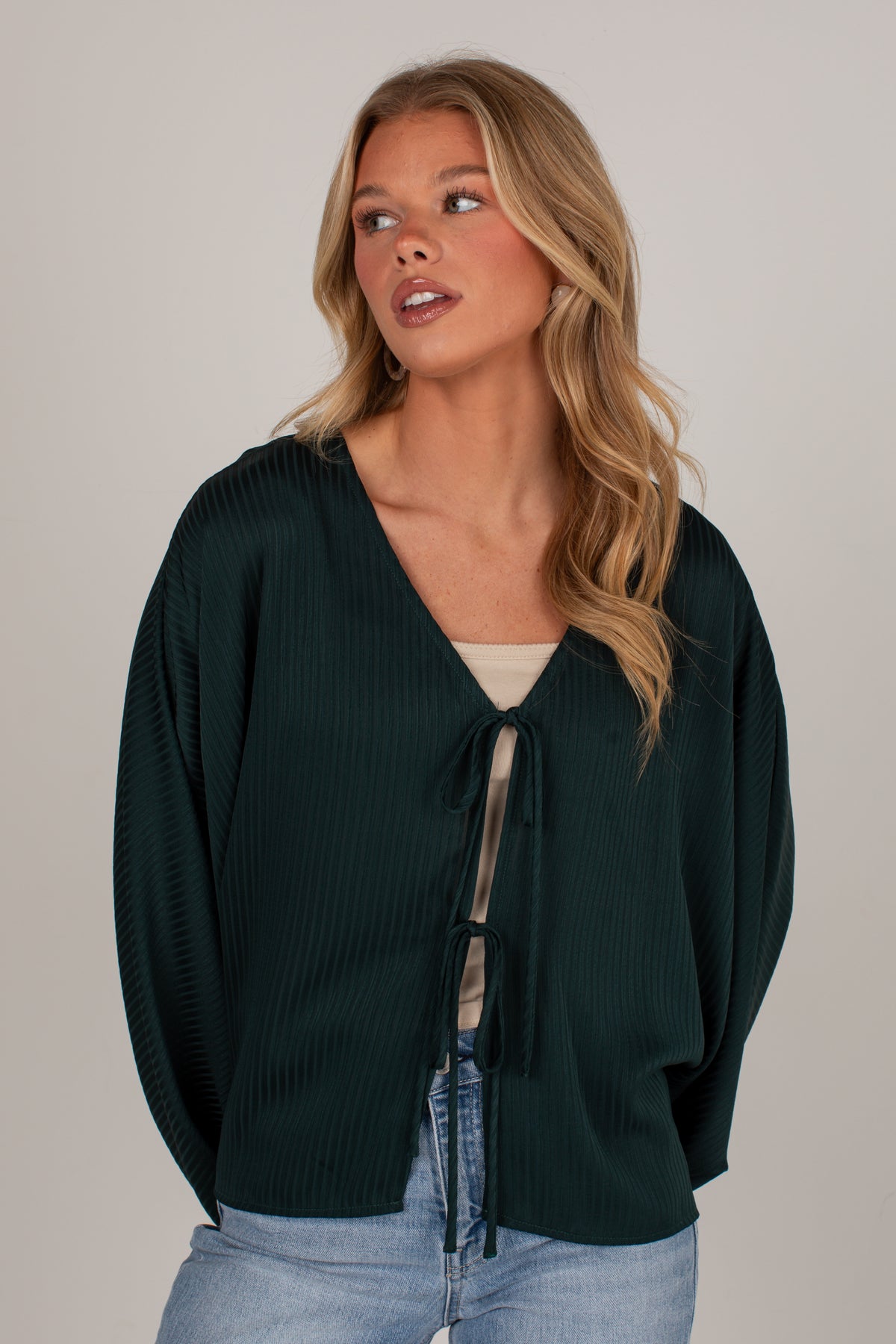 A woman wearing a dark forest green ribbed knit cardigan with a tie-front detail, styled over a beige top and light wash jeans, against a solid light gray background.
