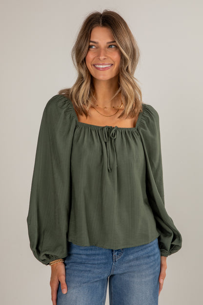 Woman wearing an olive green blouse with long sleeves and blue jeans on a plain background