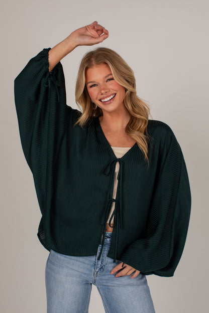 A smiling blonde woman models a dark forest green waffle-knit long-sleeve top with a tie-front closure, paired with light blue jeans, against a solid light gray studio background.