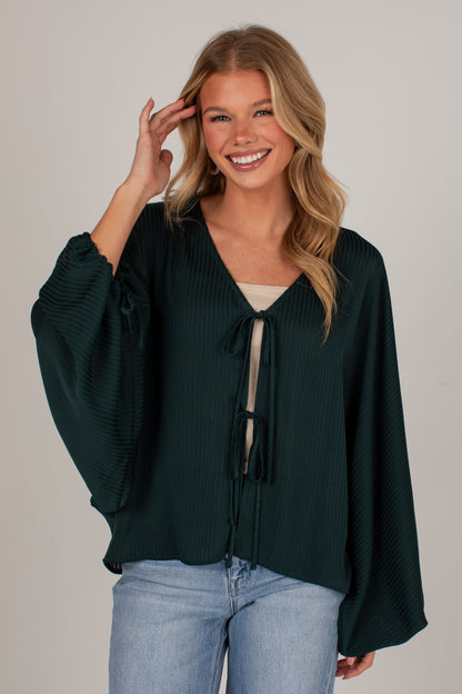 A smiling blonde woman wearing a dark forest green crinkle-textured blouse with balloon sleeves and a tie-front detail, paired with light-wash blue jeans, against a plain light gray studio background.