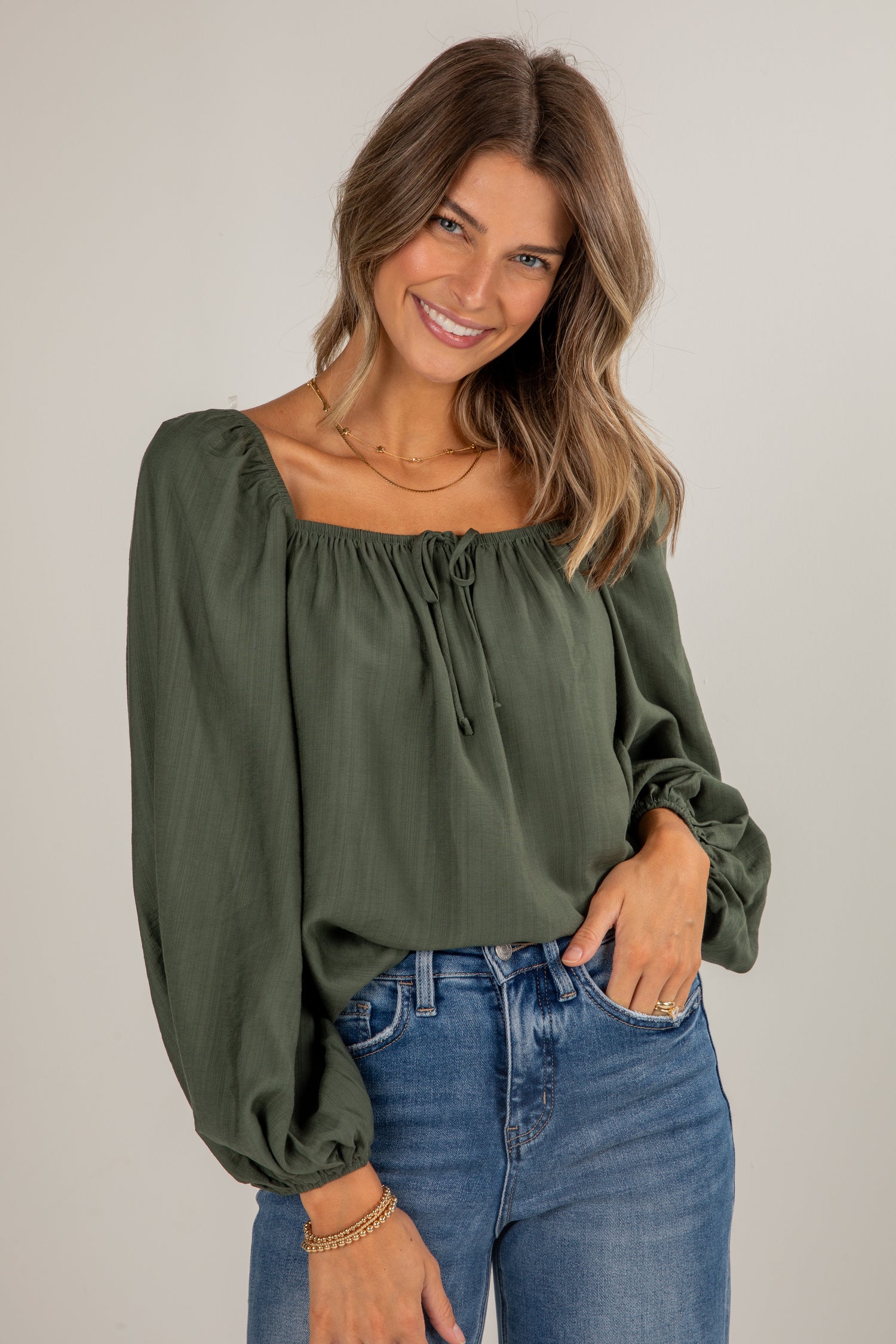Woman wearing an olive green blouse with long sleeves and blue jeans on a plain background