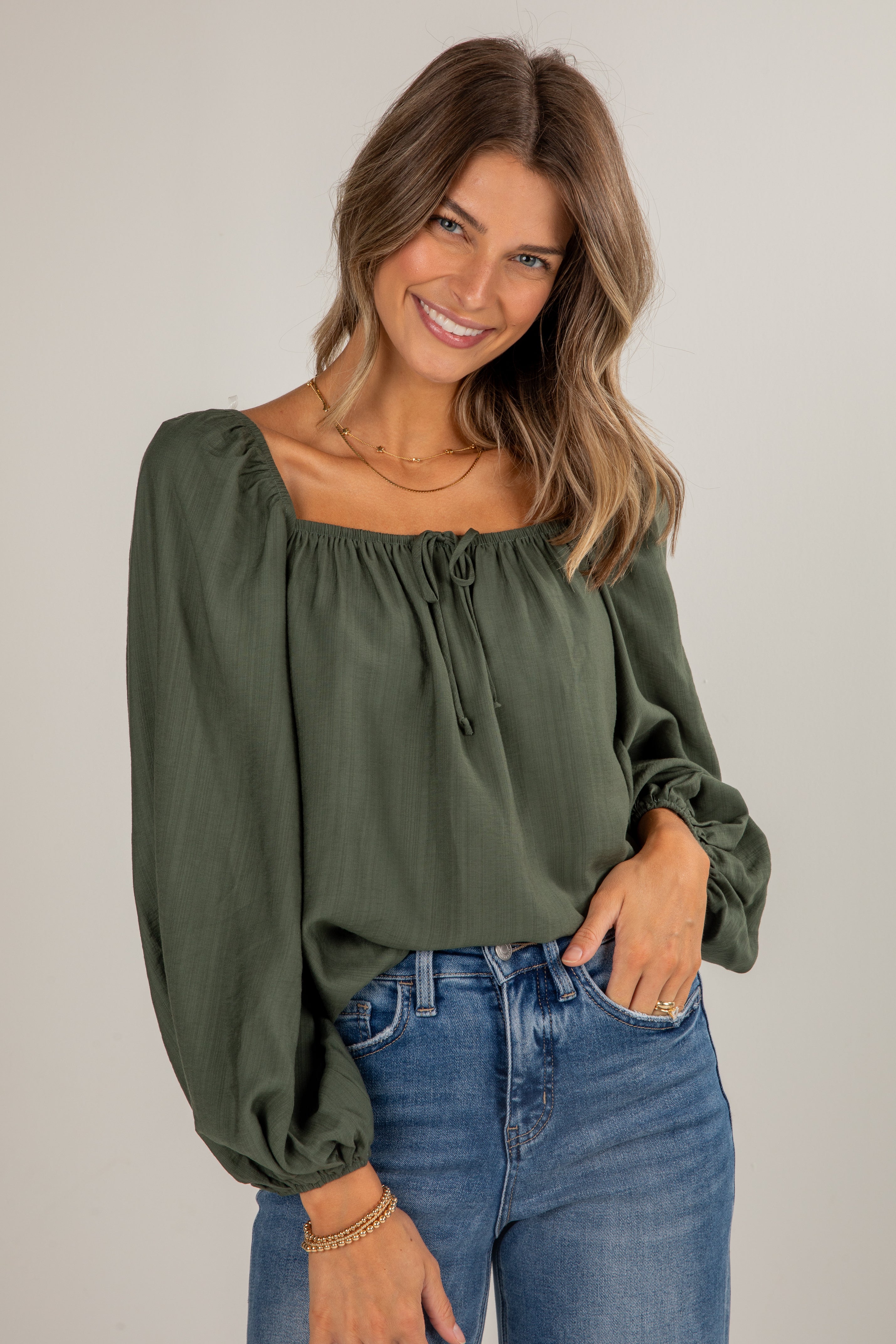 Woman wearing an olive green blouse with long sleeves and blue jeans on a plain background