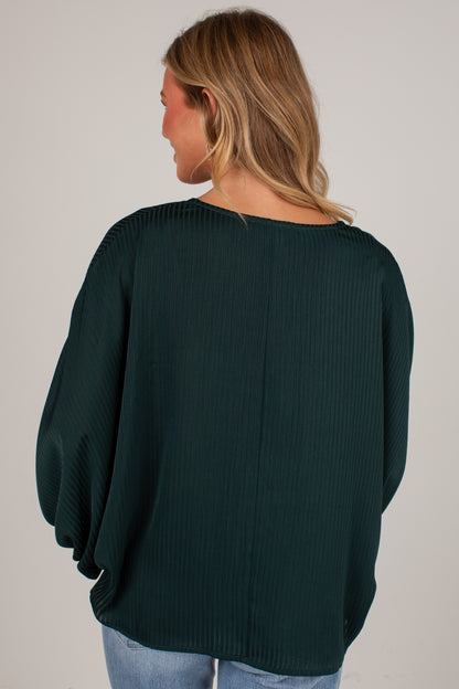 A woman viewed from behind wearing a dark forest green ribbed knit top with balloon sleeves, paired with light blue jeans, against a plain light gray background.