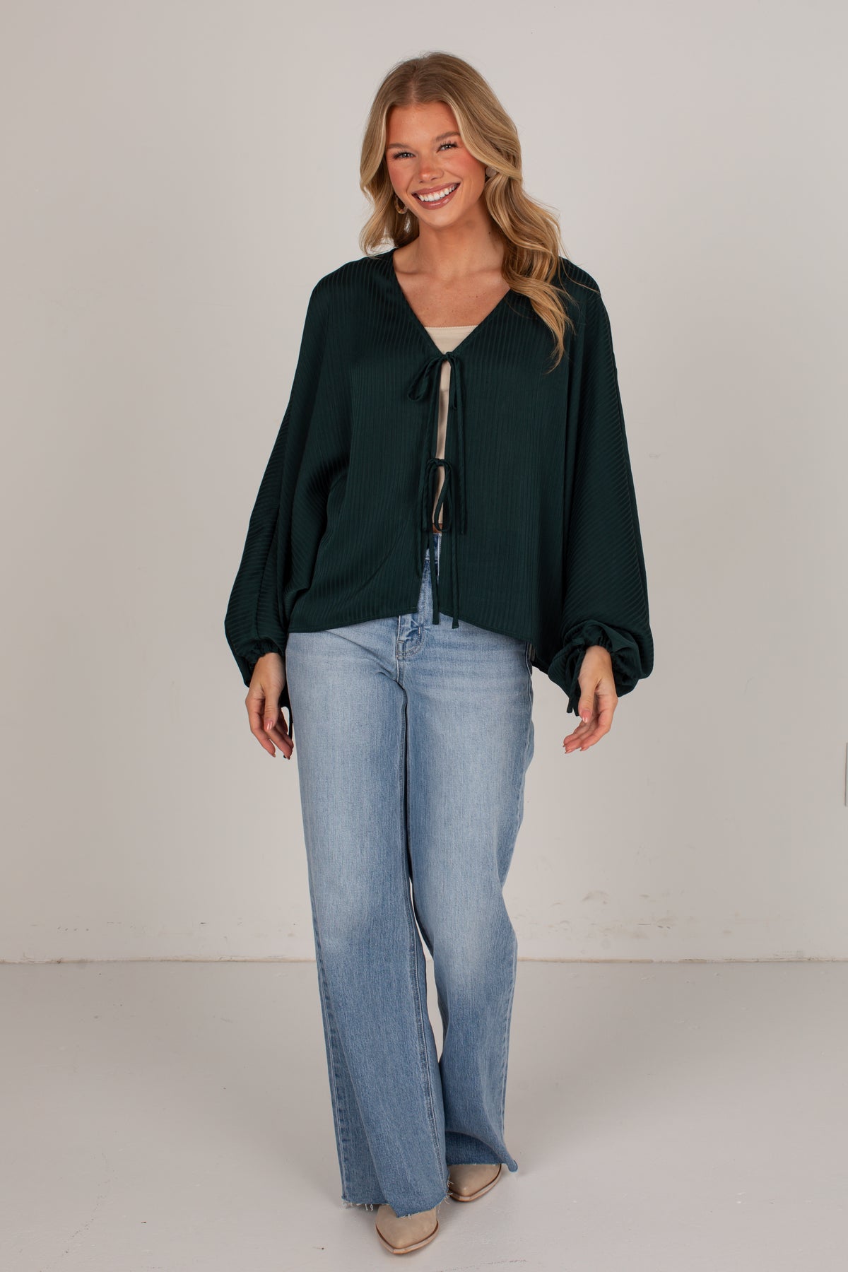 A woman modeling a dark forest green long-sleeve blouse with a tie-front detail, paired with light-wash wide-leg jeans, standing against a plain off-white studio background.