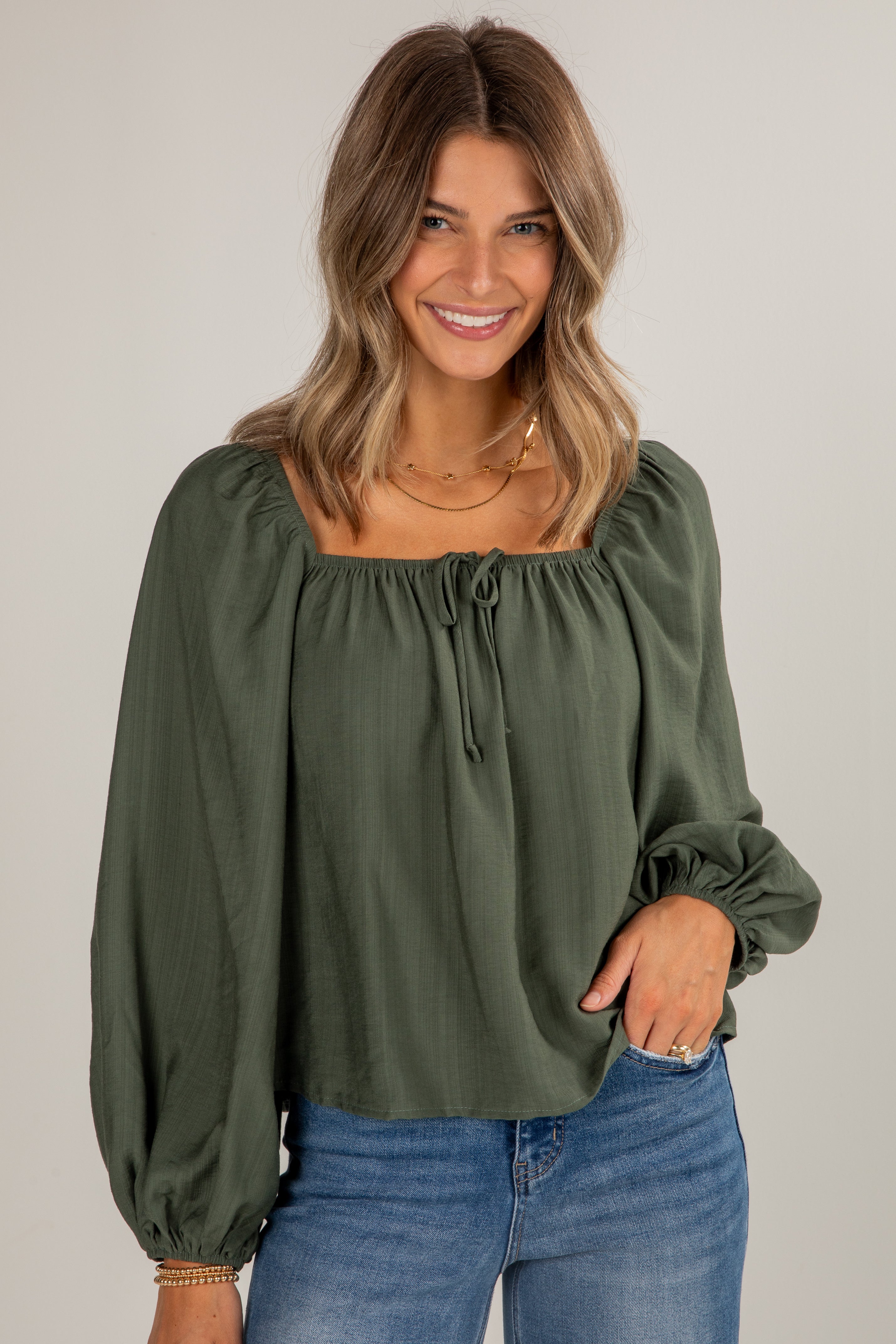 Woman wearing an olive green blouse with long sleeves and blue jeans on a plain background