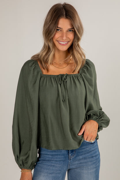 Woman wearing an olive green blouse with long sleeves and blue jeans on a plain background