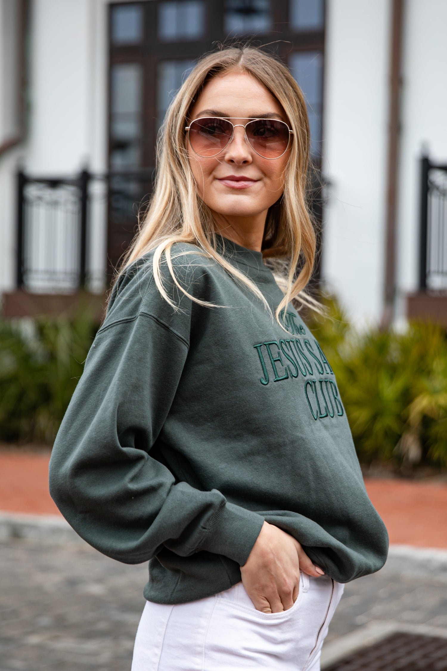 Woman wearing a green sweatshirt with text, sunglasses, and white pants outdoors.