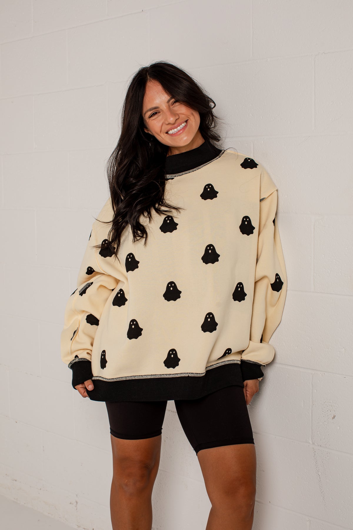 A woman wearing a cream-colored crewneck sweatshirt with black ghost patterns and black ribbed trim, paired with black biker shorts, standing against a white brick wall.