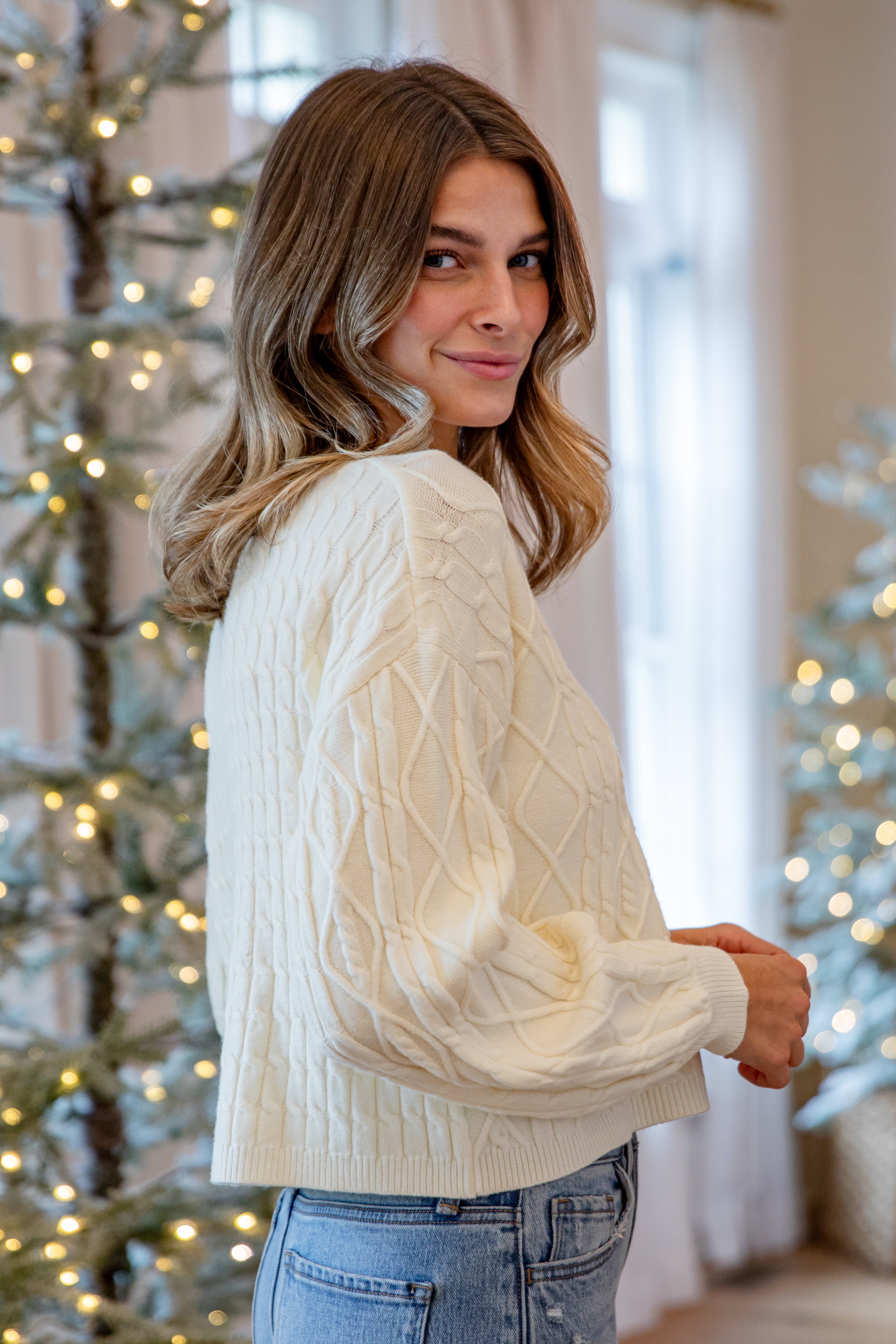 Woman wearing a white cable knit sweater in a festive indoor setting with Christmas trees.