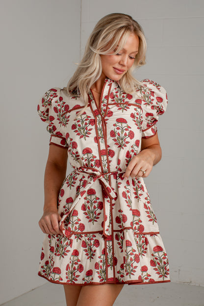 A woman modeling a short cream dress with a red and green floral print, featuring puff sleeves, a tie-neck collar, and a matching waist tie, set against a light gray wall.
