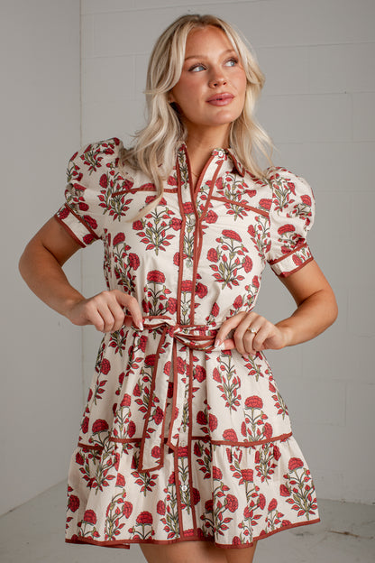 A woman modeling a short-sleeve cream dress with a red and green floral print and a matching tie belt, set against a light gray paneled wall.