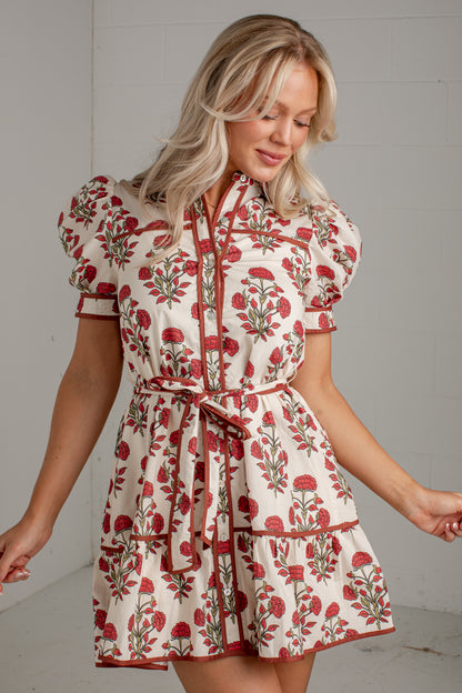 A woman wearing a short cream dress with a red and green floral print, featuring puff sleeves, a tie waist belt, and tiered skirt, standing against a light gray wall.