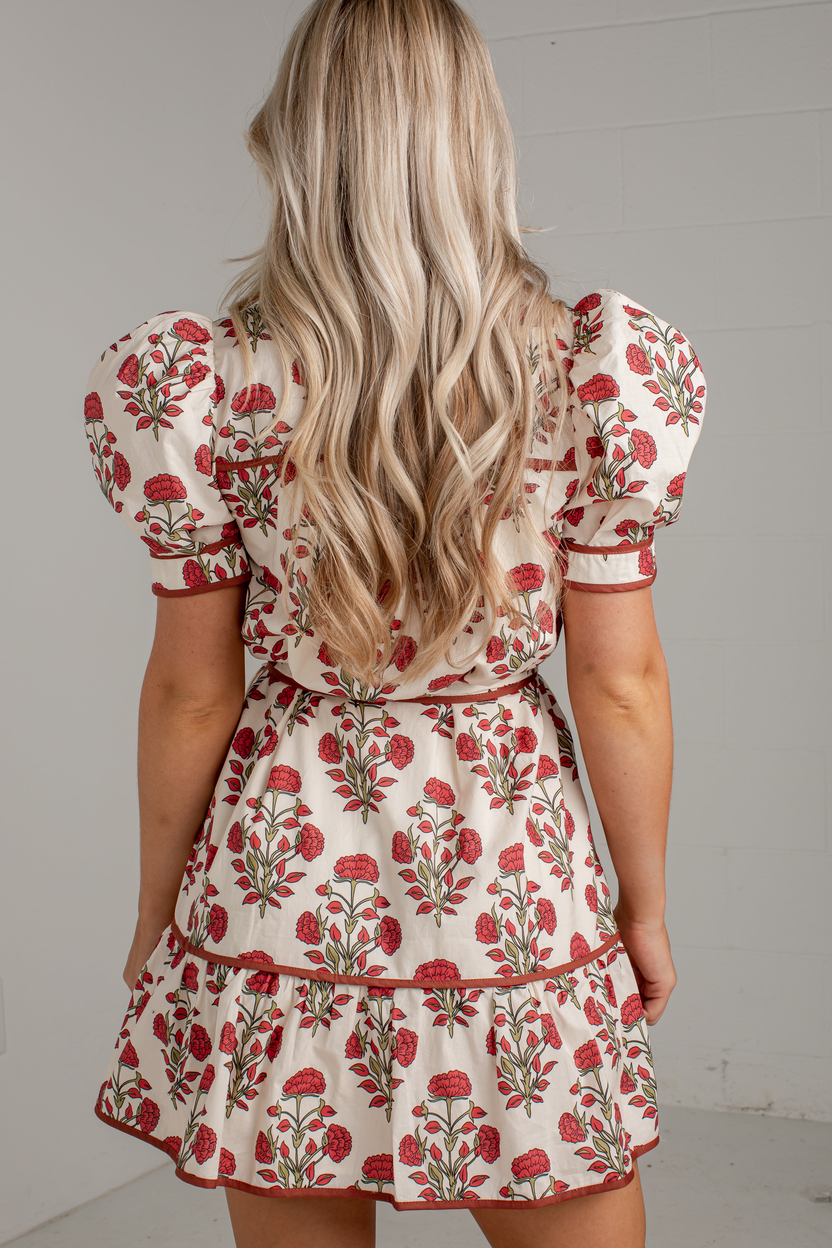 A woman viewed from behind wearing a cream-colored puff-sleeve dress with a red and green floral print, set against a plain light gray wall.