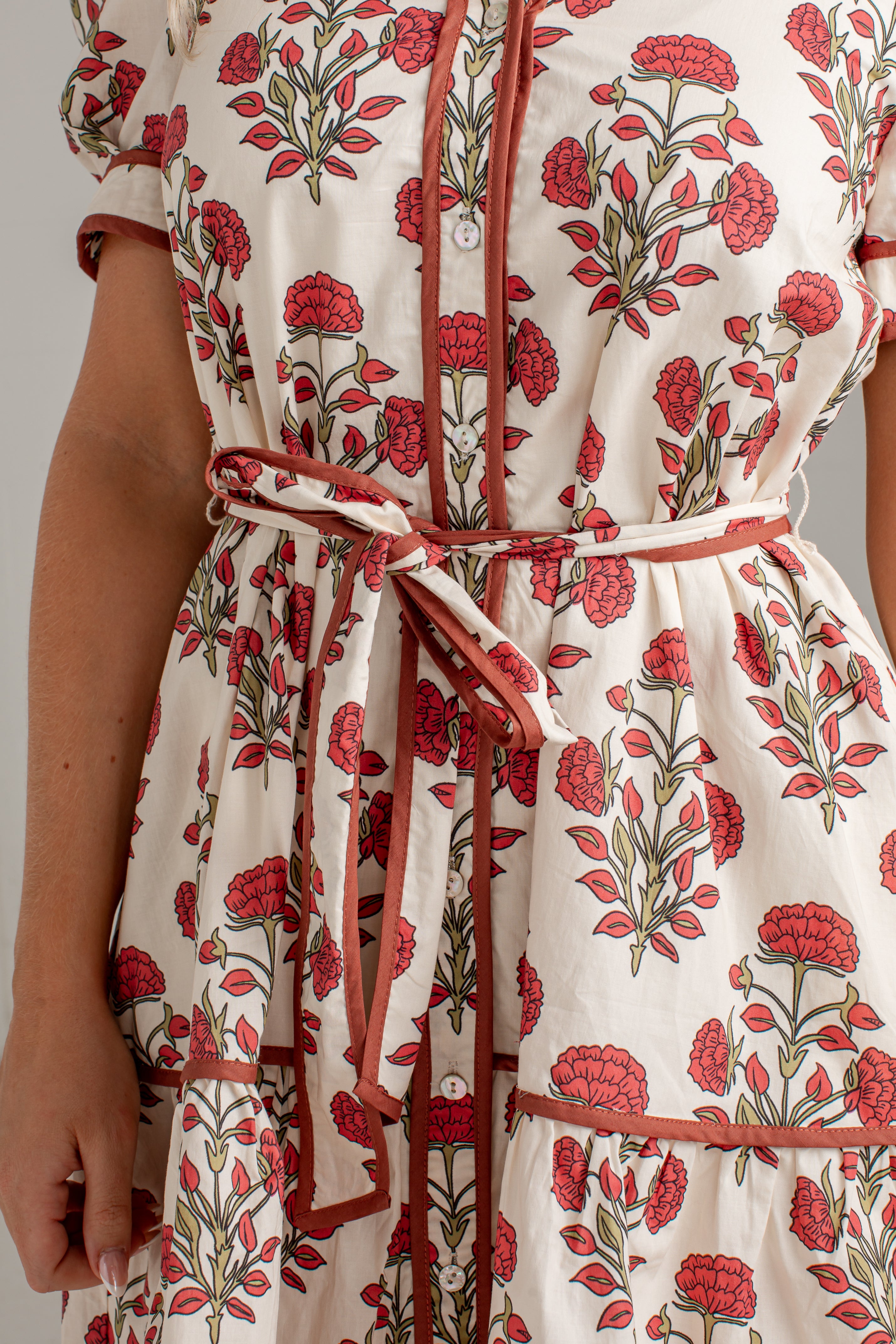 Close-up of a cream-colored dress with a red and green floral print, featuring a tie waist belt and red trim.