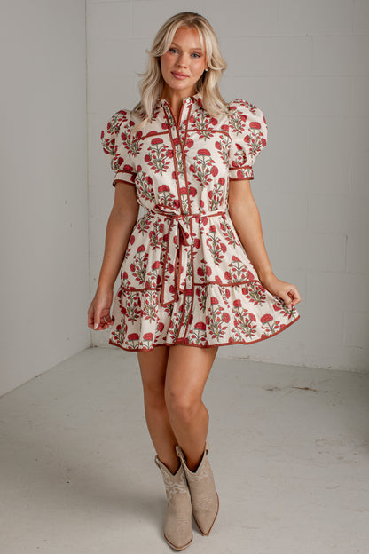 A woman modeling a short puff-sleeve dress with a red and green floral print on a cream background, paired with beige cowboy boots, in a studio setting.