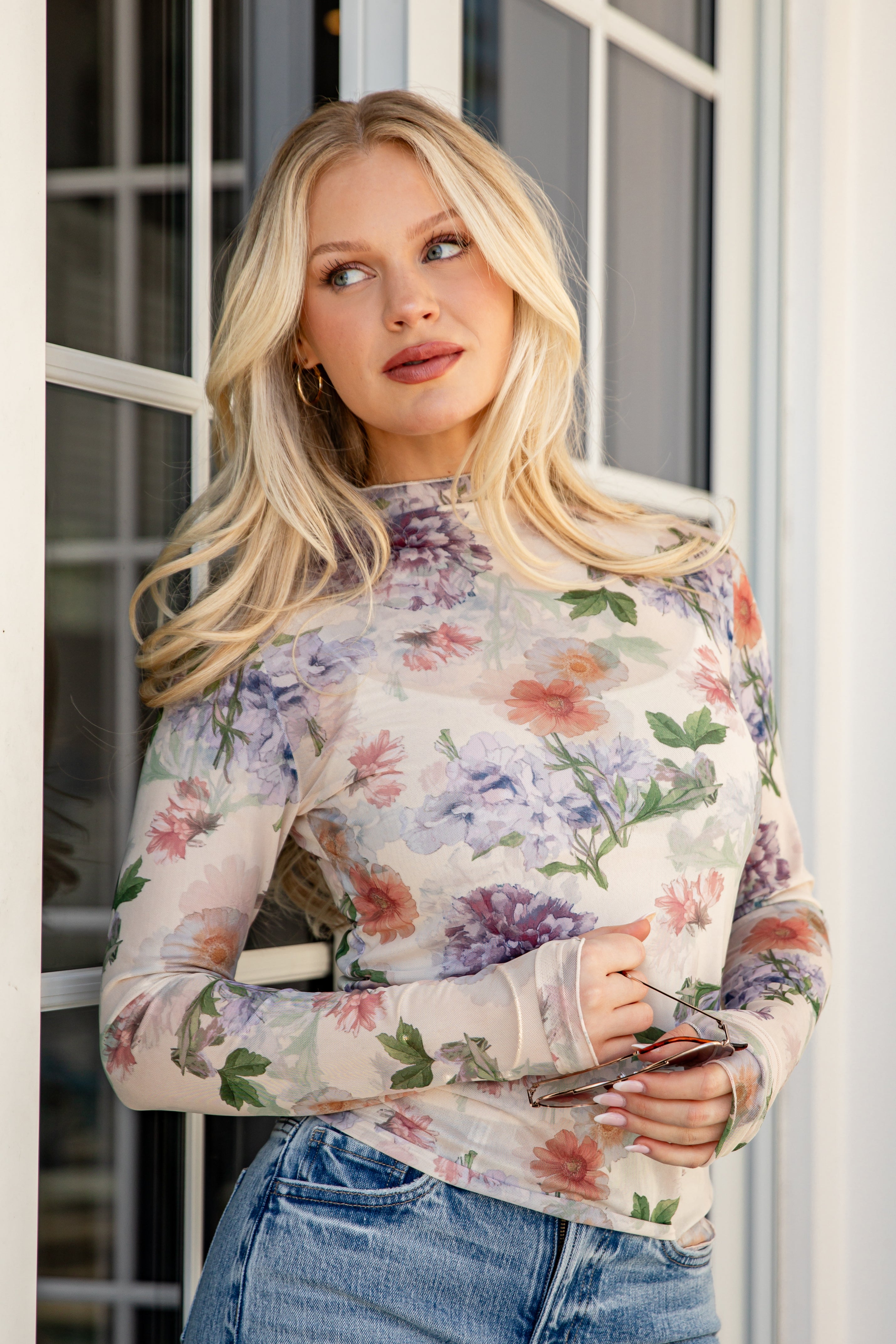 Woman wearing a floral long-sleeve top and jeans standing indoors.