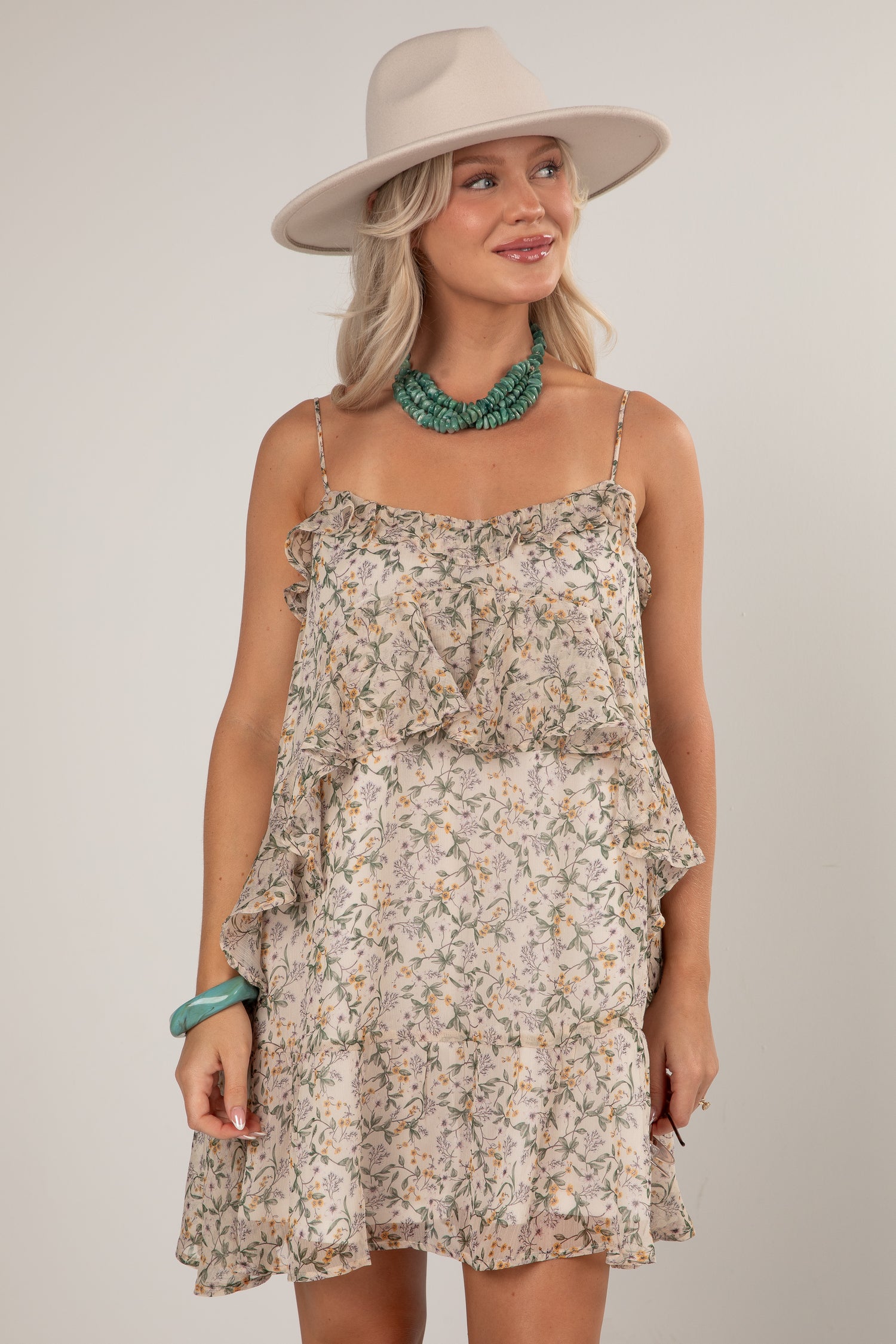A woman wearing a ruffled spaghetti-strap mini dress with a beige and green floral print, styled with a wide-brimmed beige hat, a chunky green statement necklace, and a green bracelet, set against a plain light gray studio background.