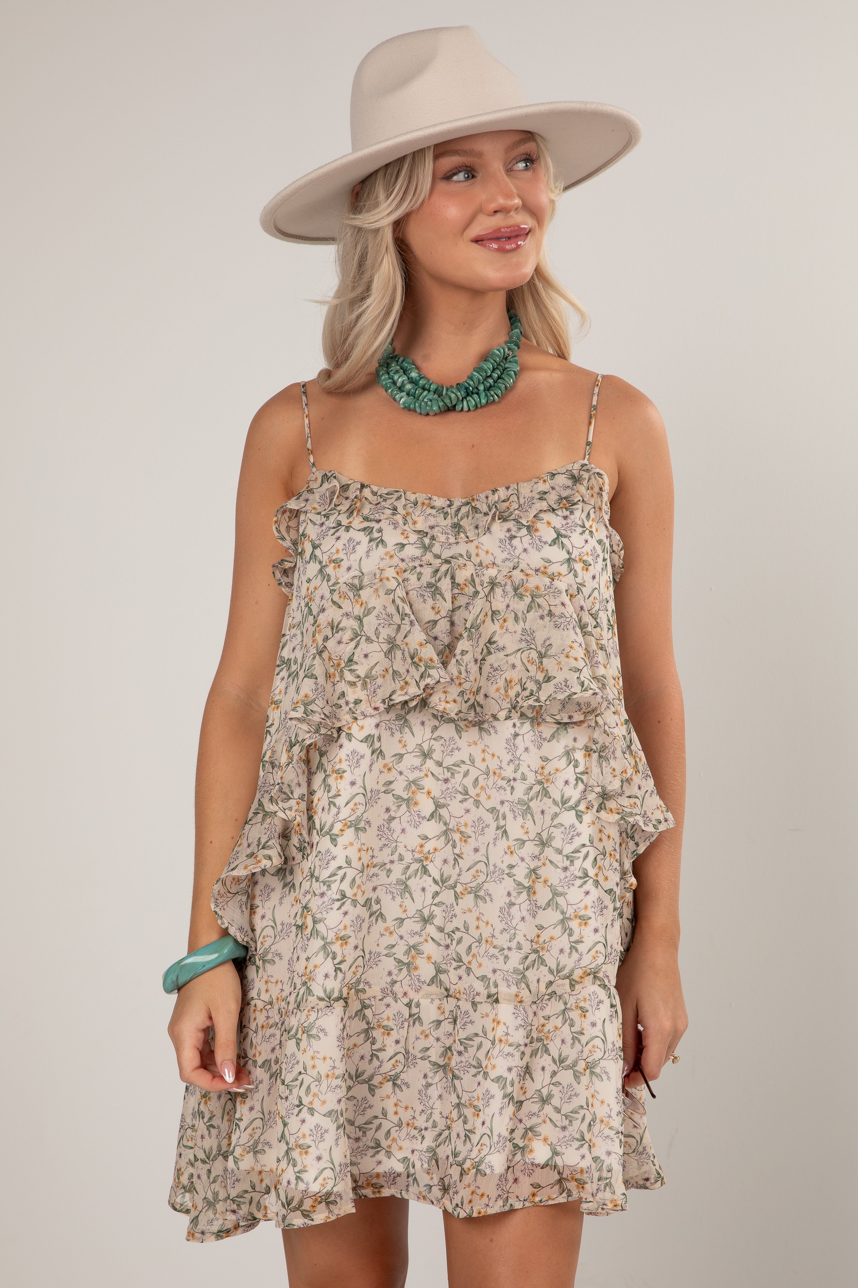 A woman wearing a ruffled spaghetti-strap mini dress with a beige and green floral print, styled with a wide-brimmed beige hat, a chunky green statement necklace, and a green bracelet, set against a plain light gray studio background.