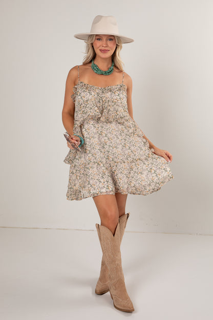 A woman modeling a short sleeveless ruffle-trimmed dress with a beige and white floral print, styled with a cream wide-brim hat, turquoise necklace, and tan cowboy boots against a plain light gray studio background.