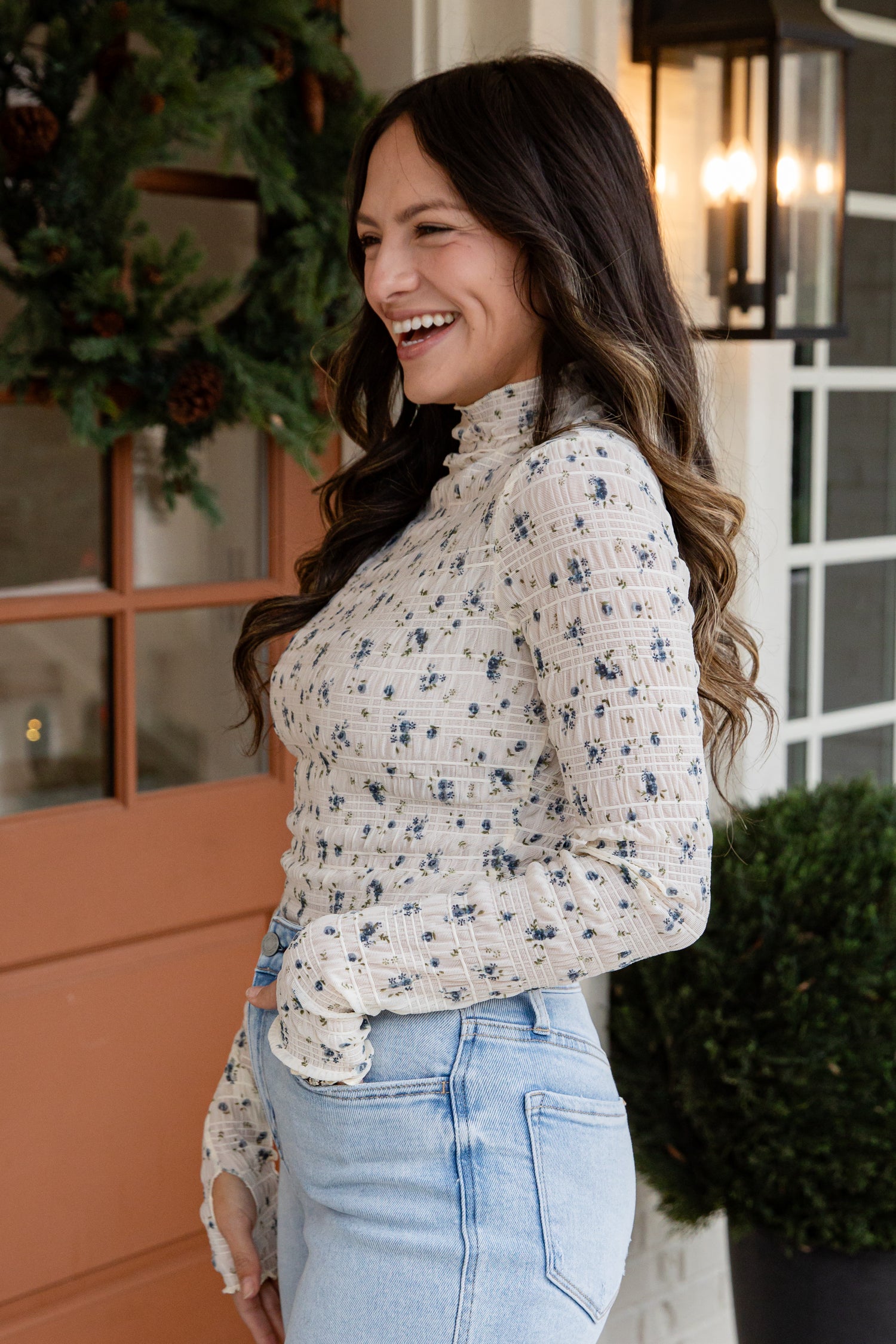 Woman wearing a floral blouse and jeans standing in front of a door with a wreath.