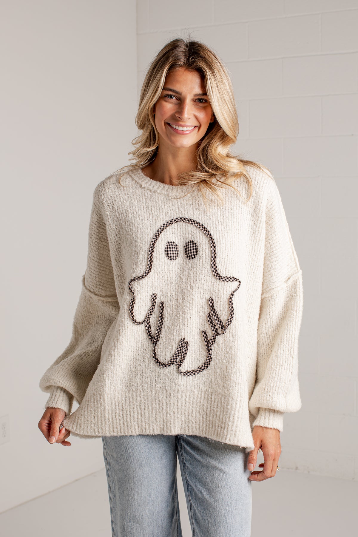 A woman wearing a cream-colored knit sweater with a large ghost motif outlined in dark brown, paired with light blue jeans, standing against a white brick wall.