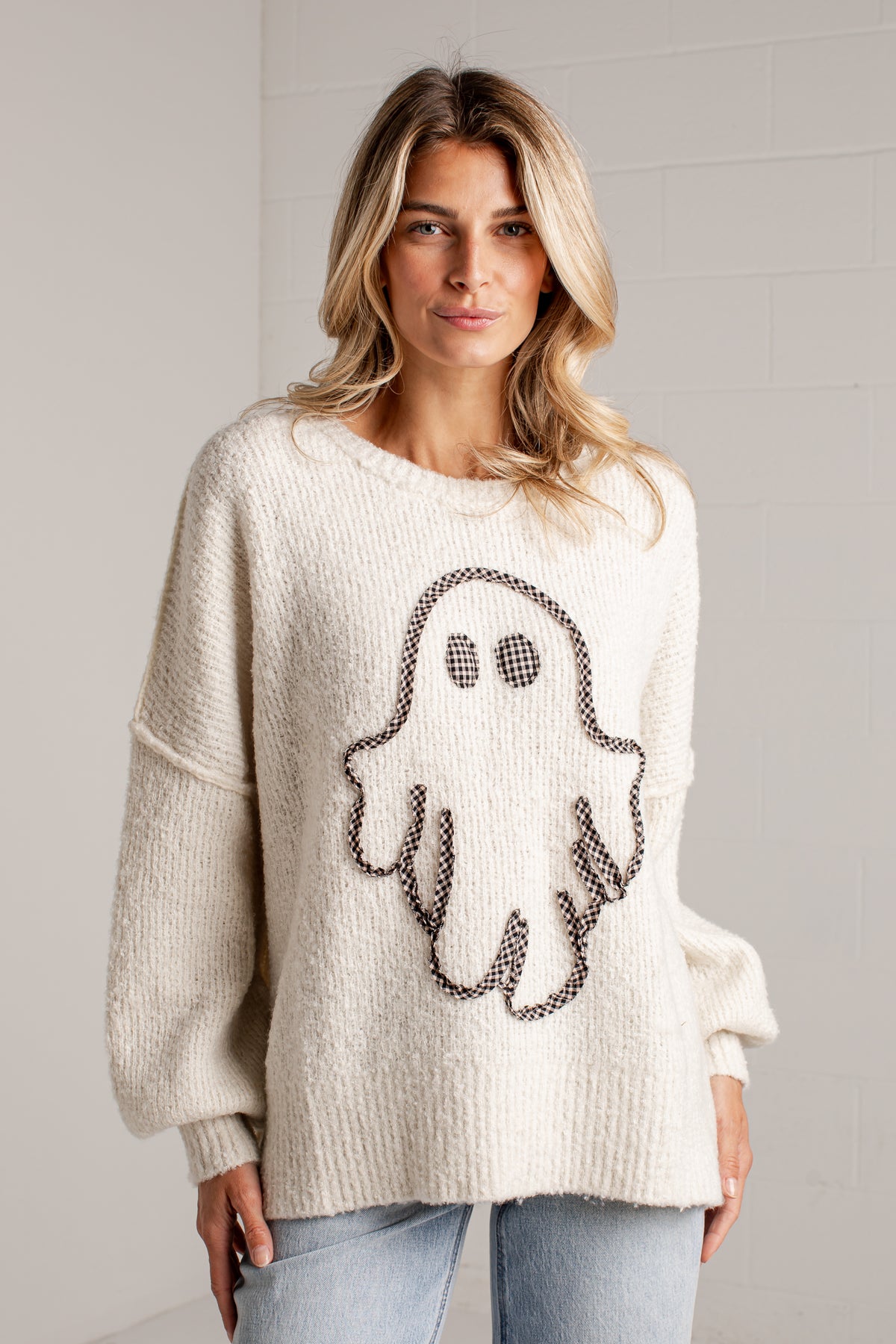 A woman wearing a cream-colored, oversized knit sweater with a black and white ghost design on the front, paired with light blue jeans, standing against a light gray brick wall.