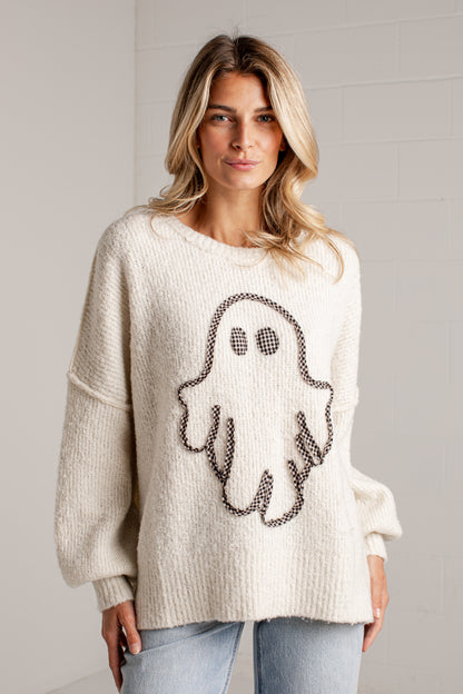 A woman wearing a cream-colored, oversized knit sweater with a black and white ghost design on the front, paired with light blue jeans, standing against a light gray brick wall.