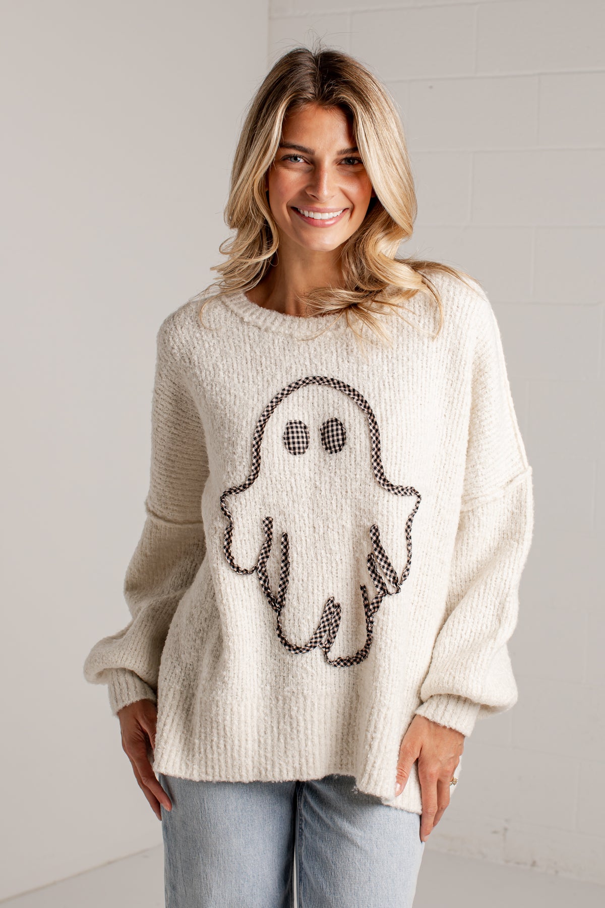 A woman wearing a cream-colored knit sweater with a black ghost graphic on the front, paired with light blue jeans, standing against a white brick wall.