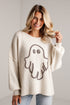 A woman wearing a cream-colored knit sweater with a large ghost graphic outlined in black on the front, paired with light blue jeans, standing against a white brick wall.