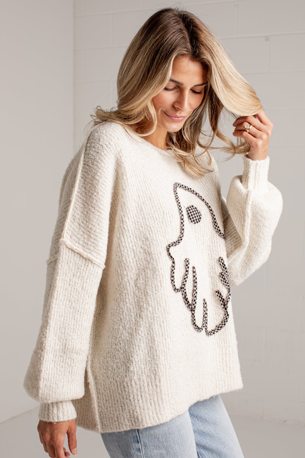 A woman wearing a cream-colored, chunky knit sweater with a ghost design outlined in black and white patterned beads on the front, paired with light blue jeans.