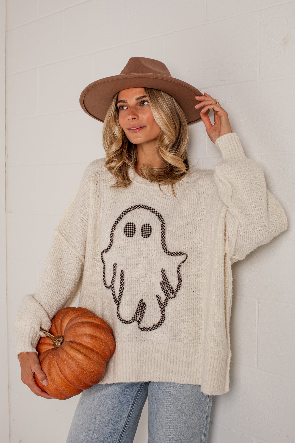 A woman wearing a cream ribbed knit sweater with a black ghost graphic, a brown wide-brim hat, and light blue jeans, holding a small orange pumpkin against a white brick wall.