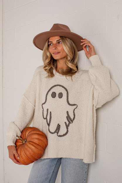 A woman wearing a cream ribbed knit sweater with a black ghost graphic, a brown wide-brim hat, and light blue jeans, holding a small orange pumpkin against a white brick wall.