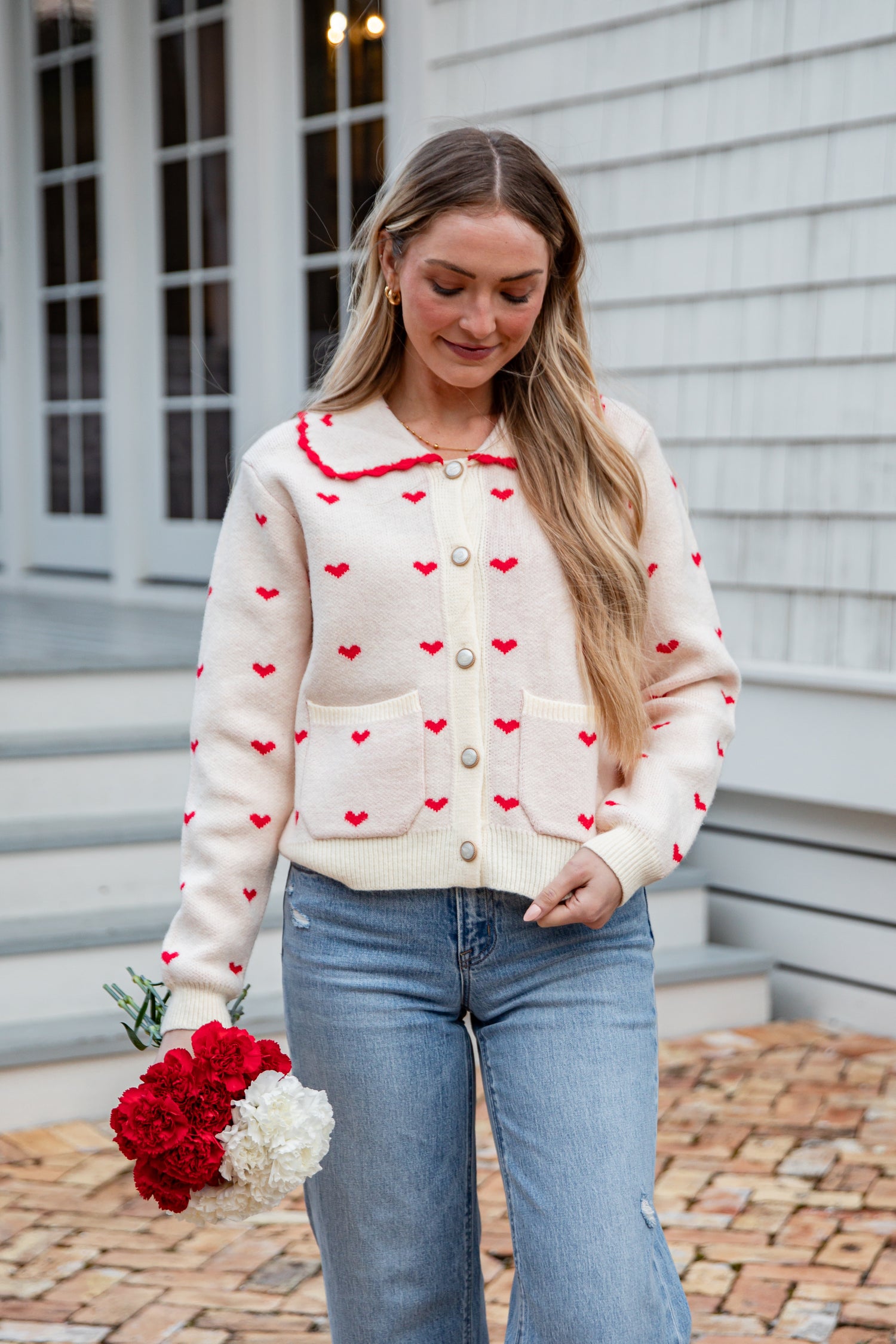 Woman wearing a cream cardigan with red heart patterns, holding flowers, standing on a brick path.