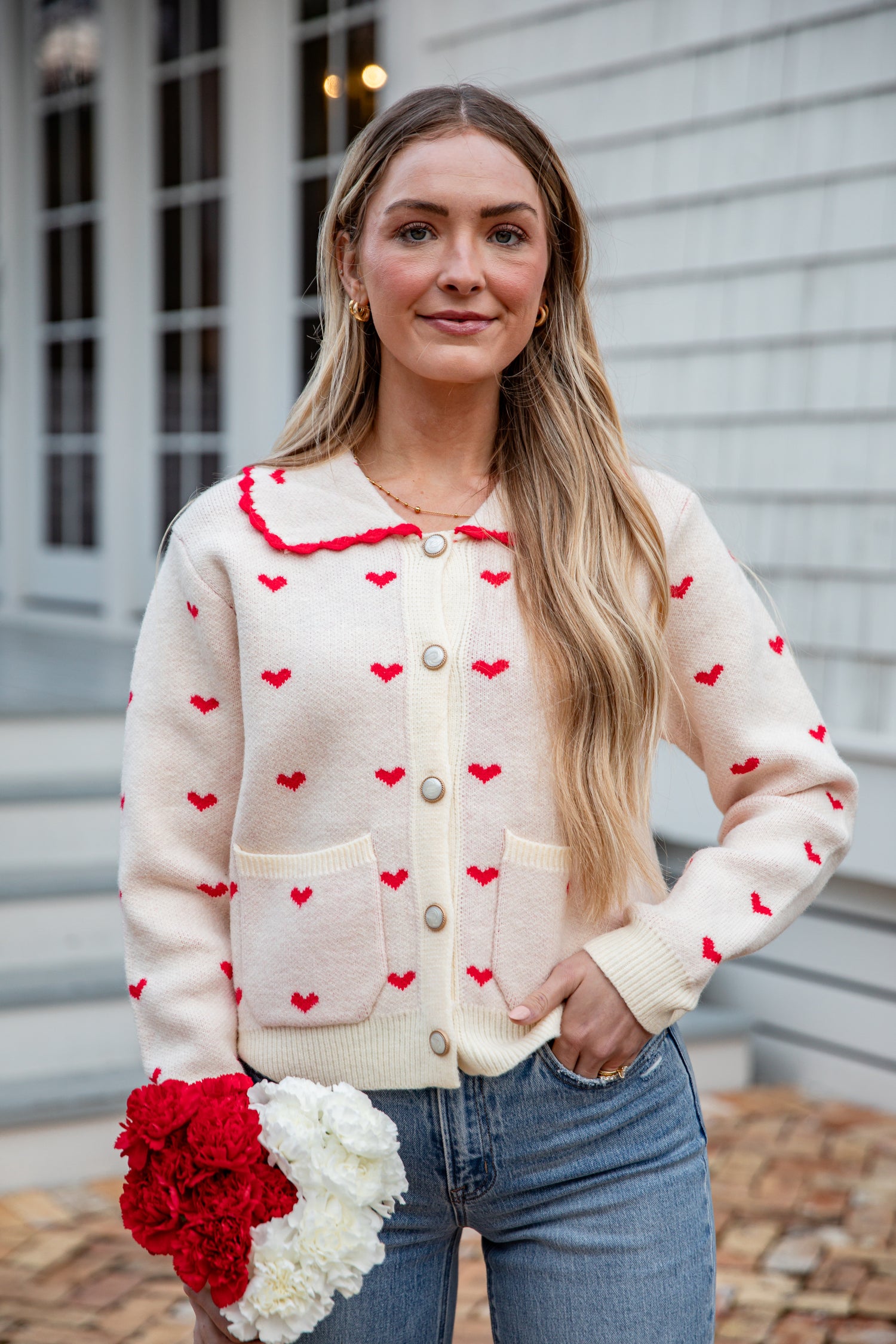 Woman wearing a cream cardigan with red heart patterns, holding flowers, standing outdoors.