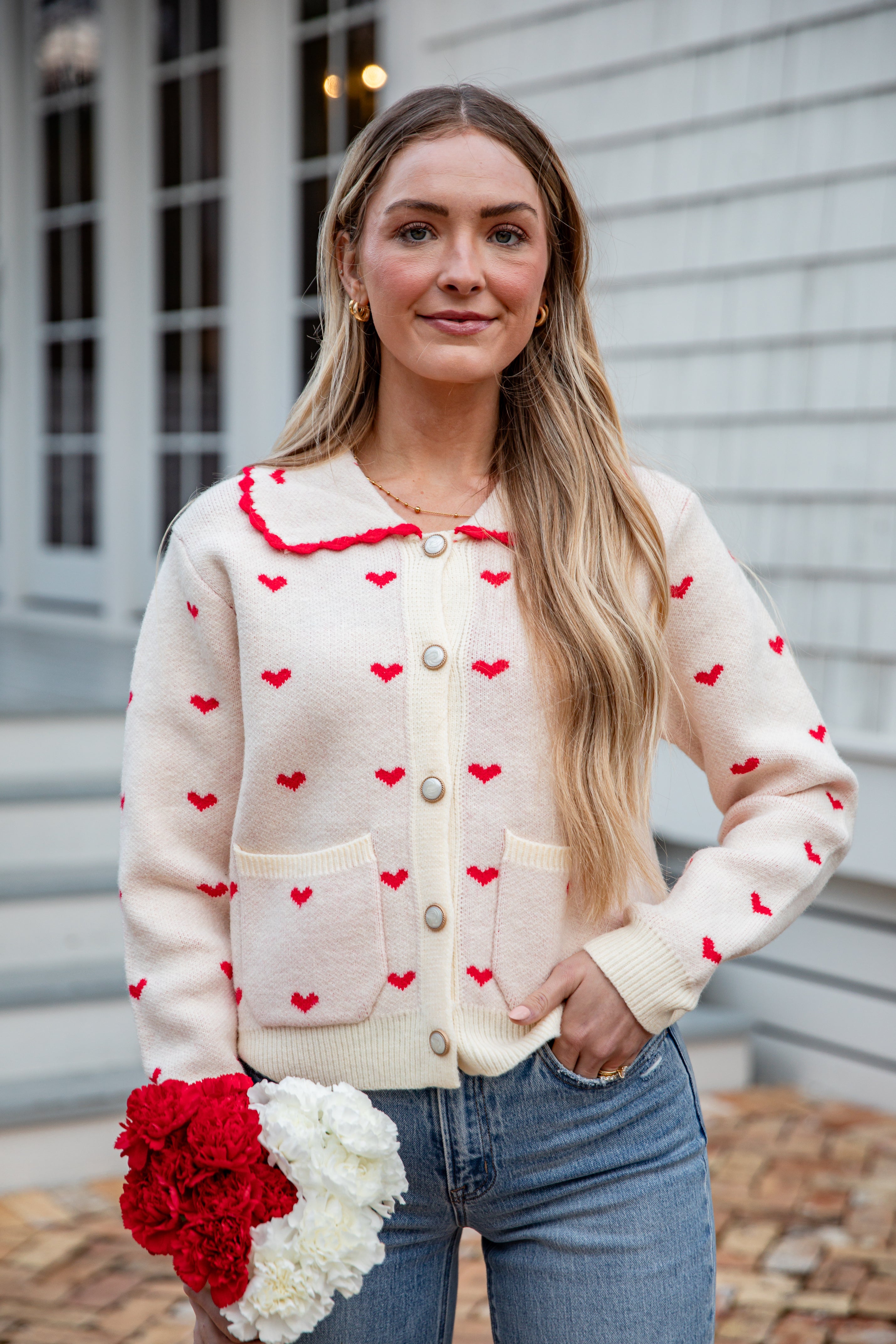 Woman wearing a cream cardigan with red heart patterns, holding flowers, standing outdoors.