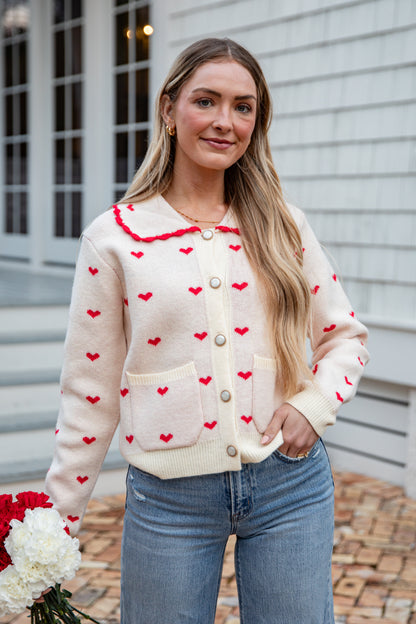 Woman wearing a cream cardigan with red heart patterns, standing outdoors.