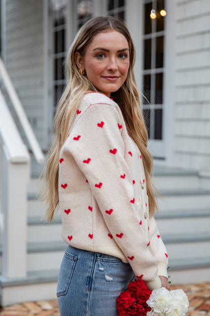 Woman wearing a cream sweater with red heart patterns, standing outdoors.