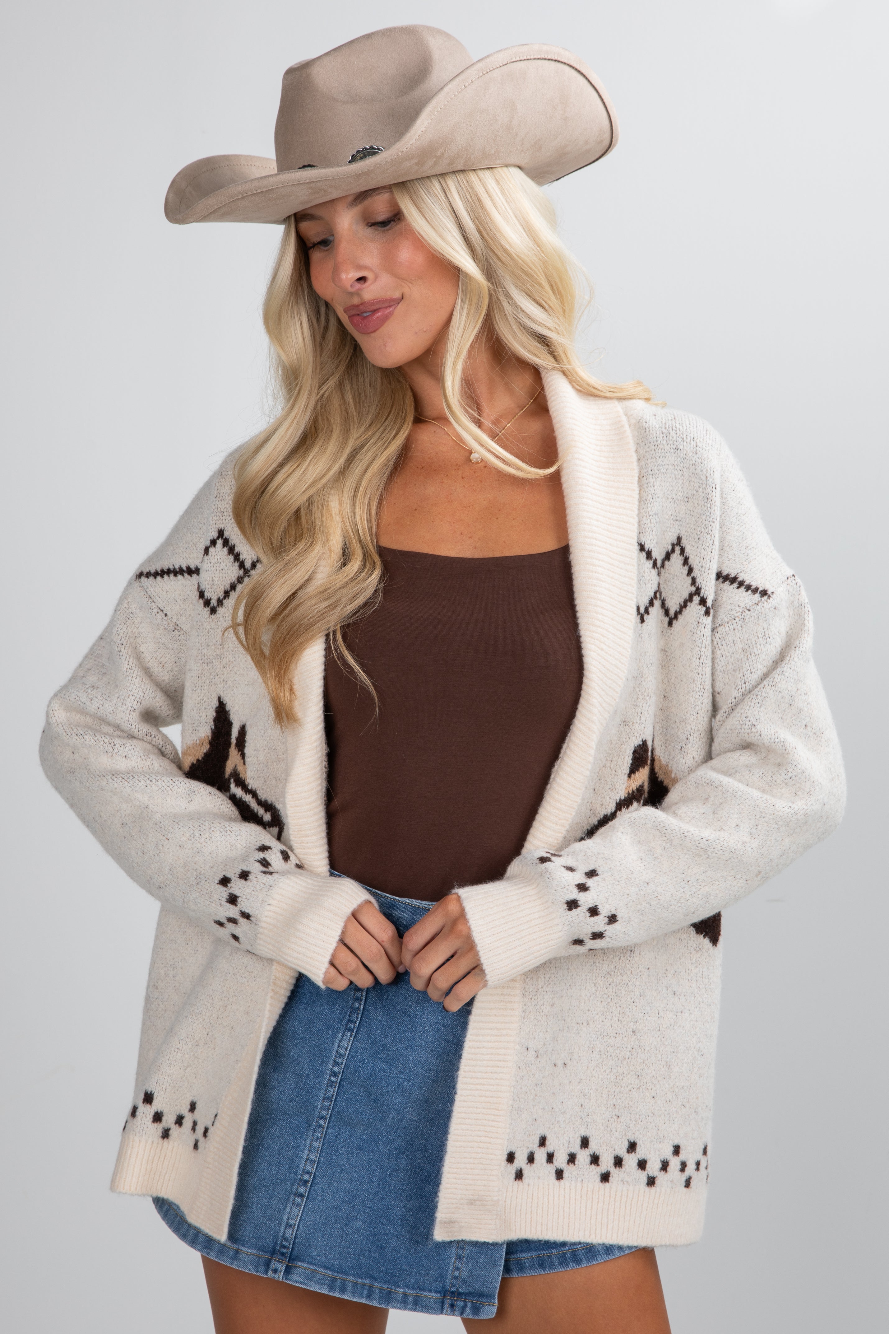 Woman wearing a beige cardigan with star patterns, brown top, and denim skirt on a gray background