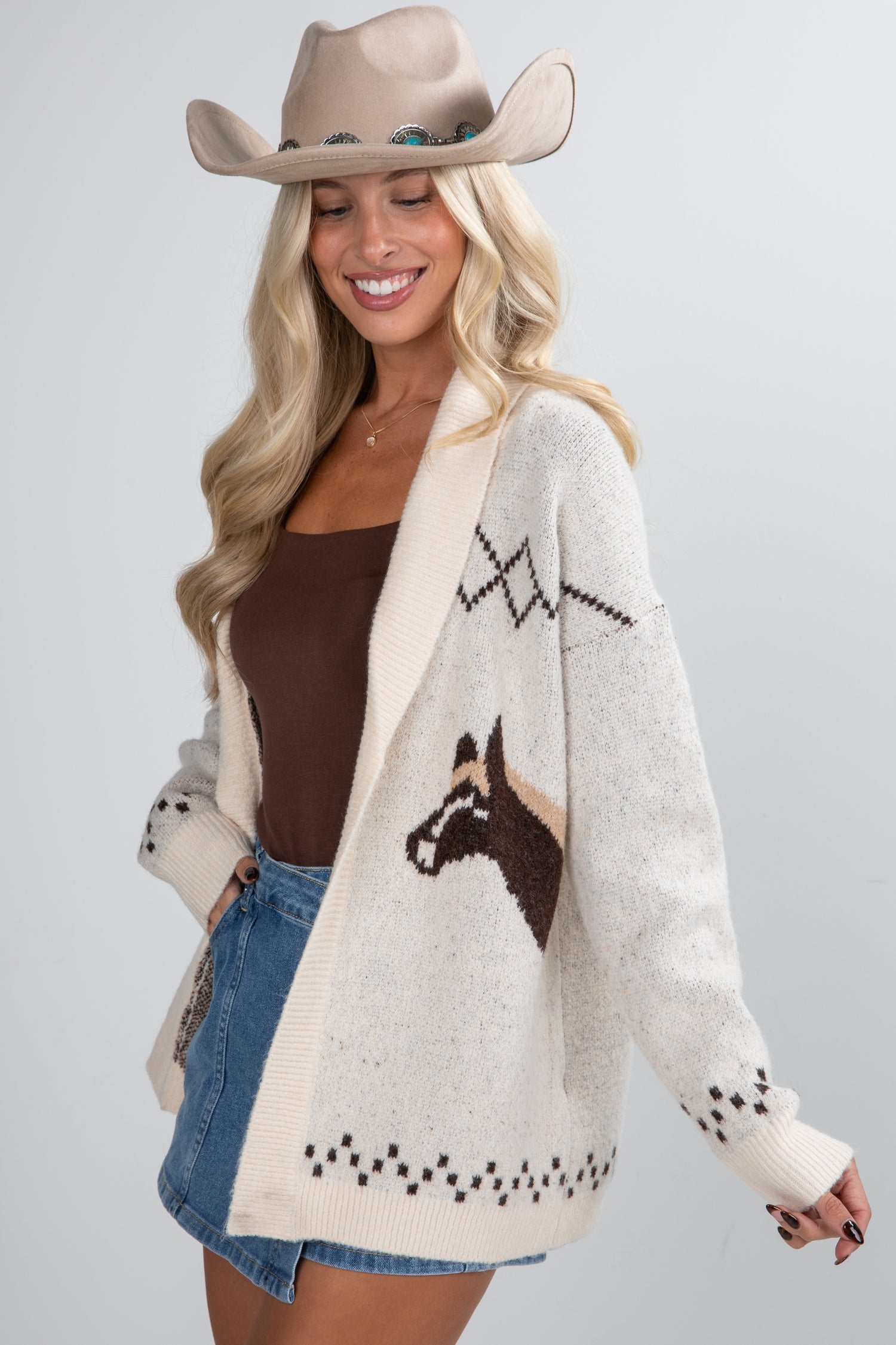 Woman wearing a cream cardigan with horse design, brown top, denim shorts, and beige cowboy hat on a light gray background.