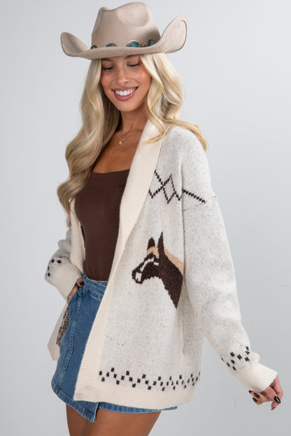 Woman wearing a cream cardigan with horse design, brown top, denim shorts, and beige cowboy hat on a light gray background.