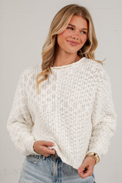 A woman wearing a chunky, textured off-white knit sweater and light-wash blue jeans, standing against a white brick wall.