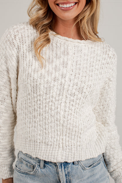 A woman wearing a chunky knit cream-colored sweater and light blue jeans, shown from the chest up against a plain light gray background.