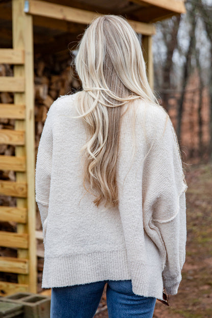 Person wearing a light gray sweater with long blonde hair, standing in a rustic outdoor setting.