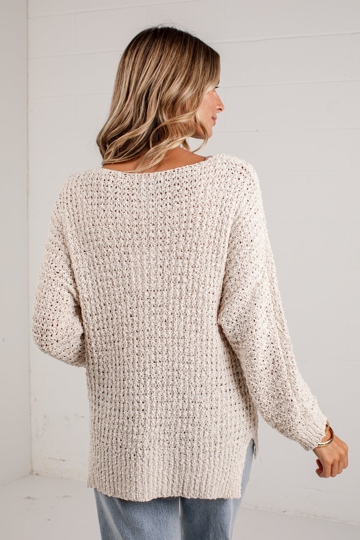 A woman wearing a chunky knit cream-colored sweater, shown from the back in a bright, white-walled room.